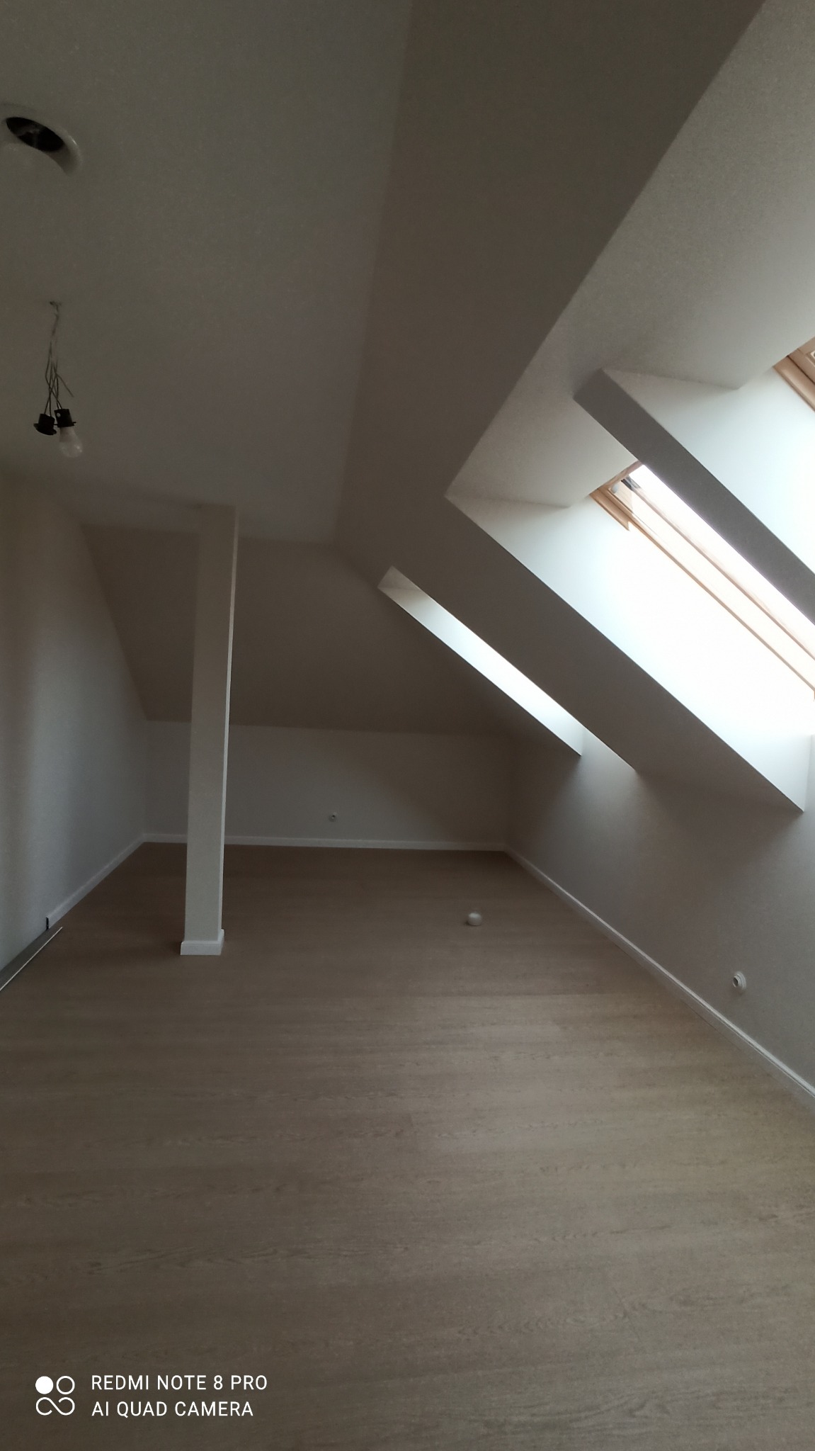 Jasne poddasze z oknami dachowymi i panelami podłogowymi. Widoczny filar i surowa żarówka. Świeżo wykończone wnętrze w neutralnych barwach.