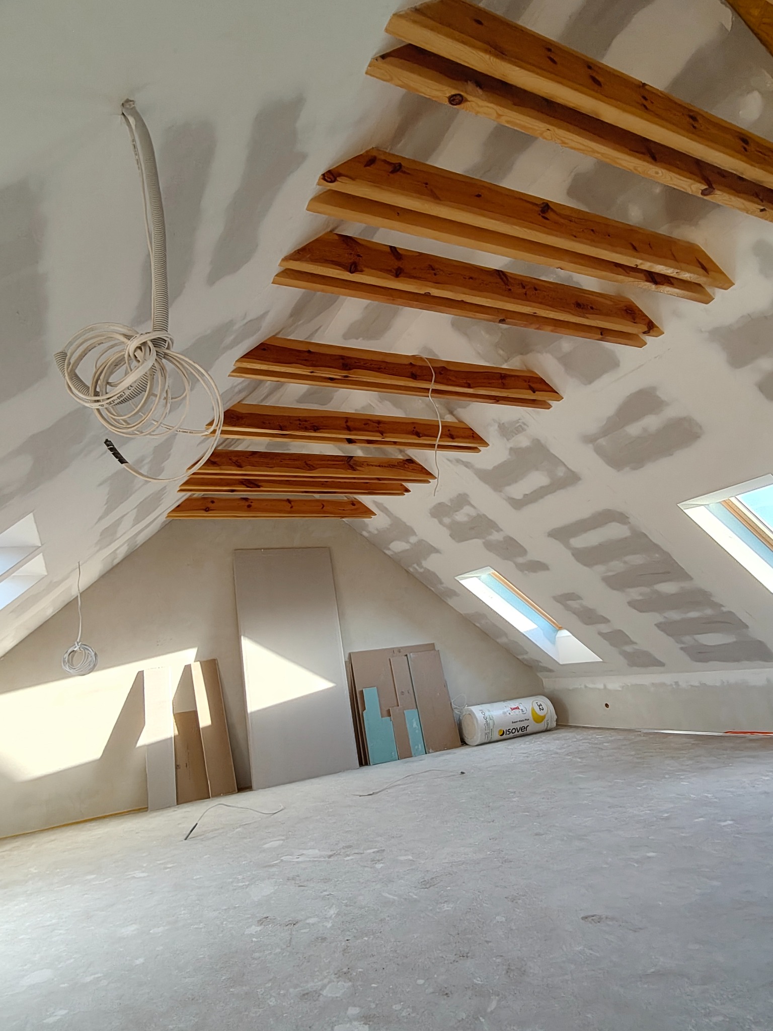 Poddasze w trakcie wykańczania: drewniane belki, płyty gipsowe, szpachlowane ściany, okna dachowe. Widoczne materiały budowlane i kable. Jasne wnętrze w surowym stanie.