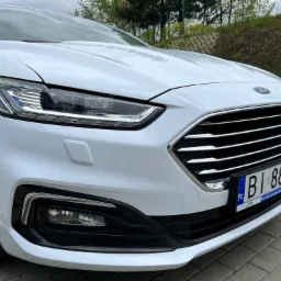 Biały samochód marki Ford z widoczną tablicą rejestracyjną na tle zieleni.