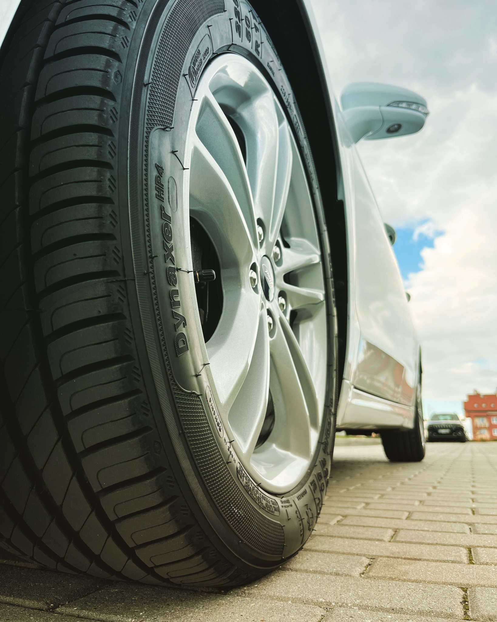 Detaliczny widok opony Dynaxer HP4 na srebrnej feldze białego samochodu, sfotografowany z niskiej perspektywy na tle kostki brukowej i błękitnego nieba z chmurami.
