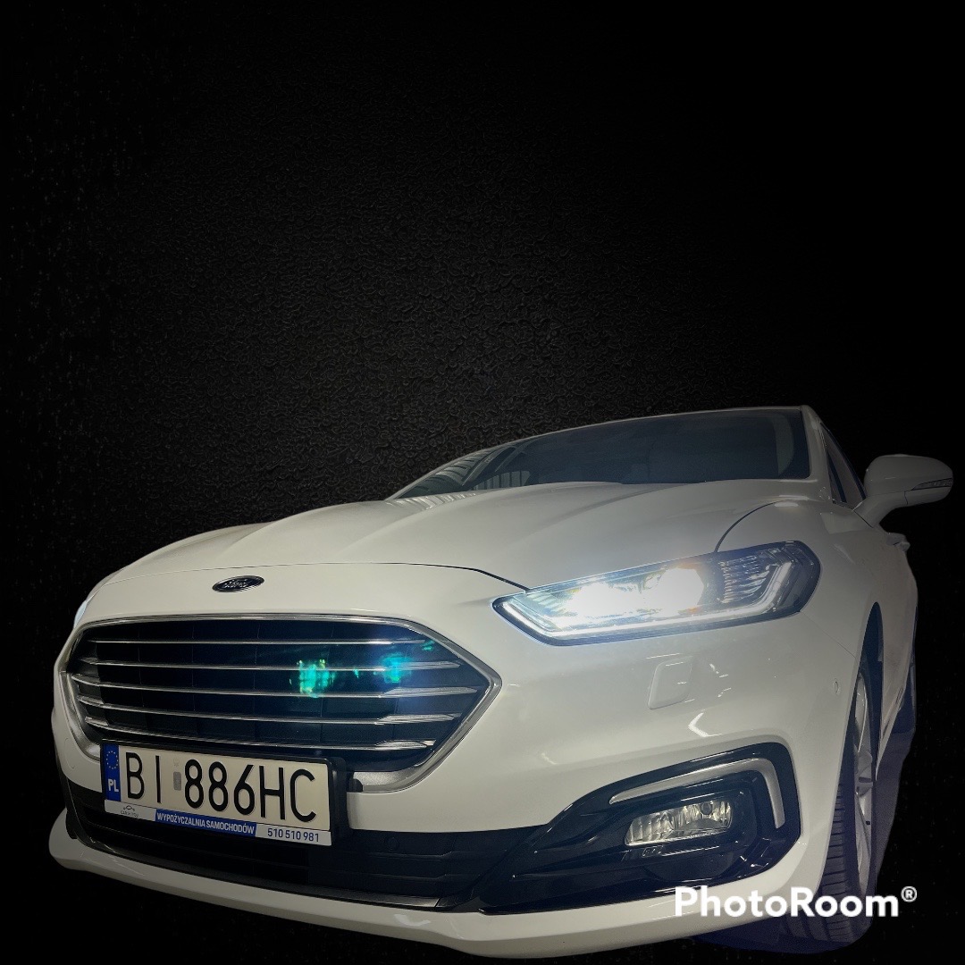 Biały samochód Ford z włączonymi światłami, widoczny z przodu, z tablicą rejestracyjną 'BI 886HC' i napisem 'Wypożyczalnia samochodów' na ramce tablicy.