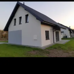 Nowoczesny dom jednorodzinny z szarą elewacją i czarnym dachem, widok z boku na tle zielonego trawnika i drzew