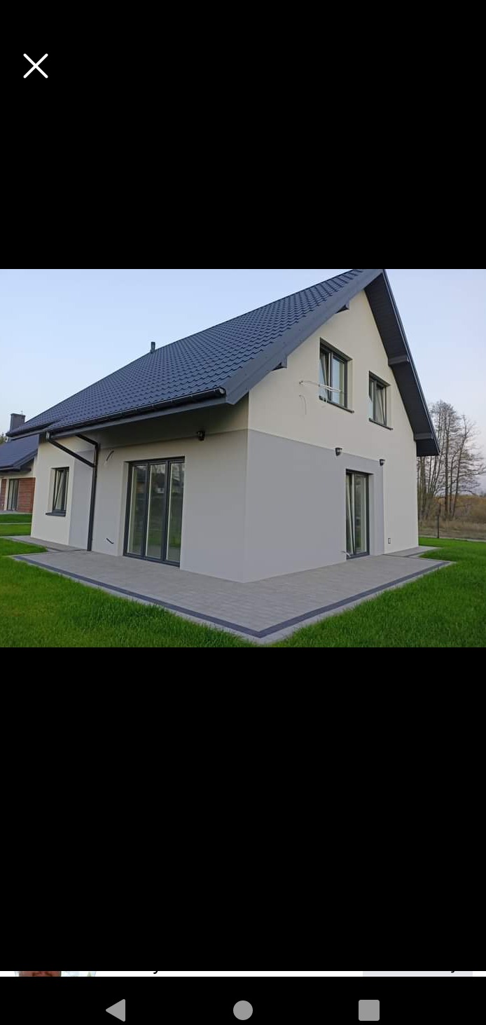 Dwukondygnacyjny dom jednorodzinny z szarą elewacją i grafitowym dachem, widok z zewnątrz na fragment fasady i przyległy chodnik z kostki brukowej.