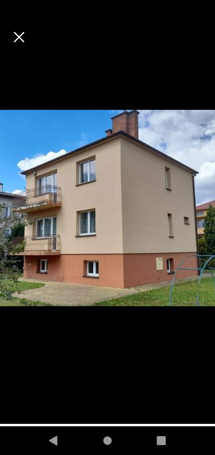 Dwukondygnacyjny budynek z balkonami po renowacji, z jasną elewacją i brązowym cokołem, widoczny z poziomu trawnika, w tle drzewa i błękitne niebo z chmurami.