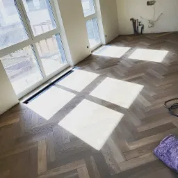 Ułożony parkiet w jodełkę francuską, z widocznymi promieniami słońca wpadającymi przez okno, ukazujący detale drewnianej podłogi i fragment instalacji na ścianie w surowym stanie.