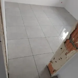 Świeżo ułożone, jasnoszare płytki podłogowe w pomieszczeniu w trakcie prac wykończeniowych, widoczny fragment surowej cegły i ślady zaprawy na ścianie.