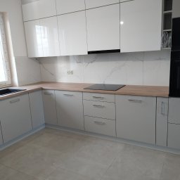 Wykończenia wnętrz - Nowoczesna kuchnia z białymi, lakierowanymi szafkami górnymi i szarymi dolnymi, drewnianym blatem, płytą indukcyjną i marmurowym panelem ściennym, widok na okno i płytki podłogowe.