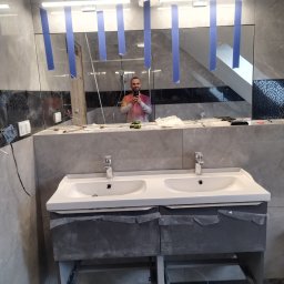 Wykończenia wnętrz - Podwójna umywalka z szarą szafką w trakcie montażu w łazience z szarymi płytkami, widoczne narzędzia i odbicie osoby robiącej zdjęcie w lustrze zabezpieczonym niebieską taśmą.