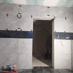 Wykończenia wnętrz - Szare płytki imitujące marmur, ułożone na ścianie z użyciem systemu poziomowania, z czarnym pasem dekoracyjnym na wysokości wzroku, widoczne butelki z napojami na podłodze i osoba w tle w przejściu.