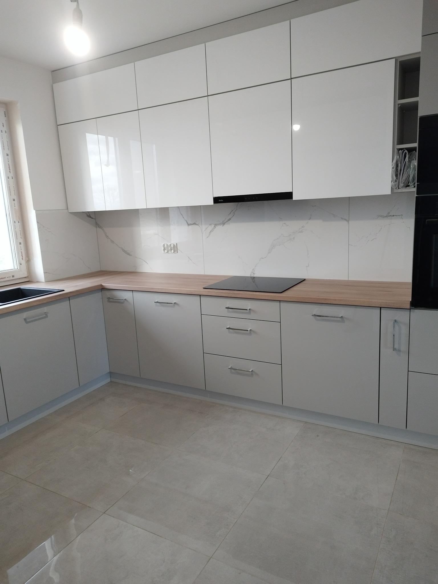 Nowoczesna kuchnia z białymi, lakierowanymi szafkami górnymi i szarymi dolnymi, drewnianym blatem, płytą indukcyjną i marmurowym panelem ściennym, widok na okno i płytki podłogowe.