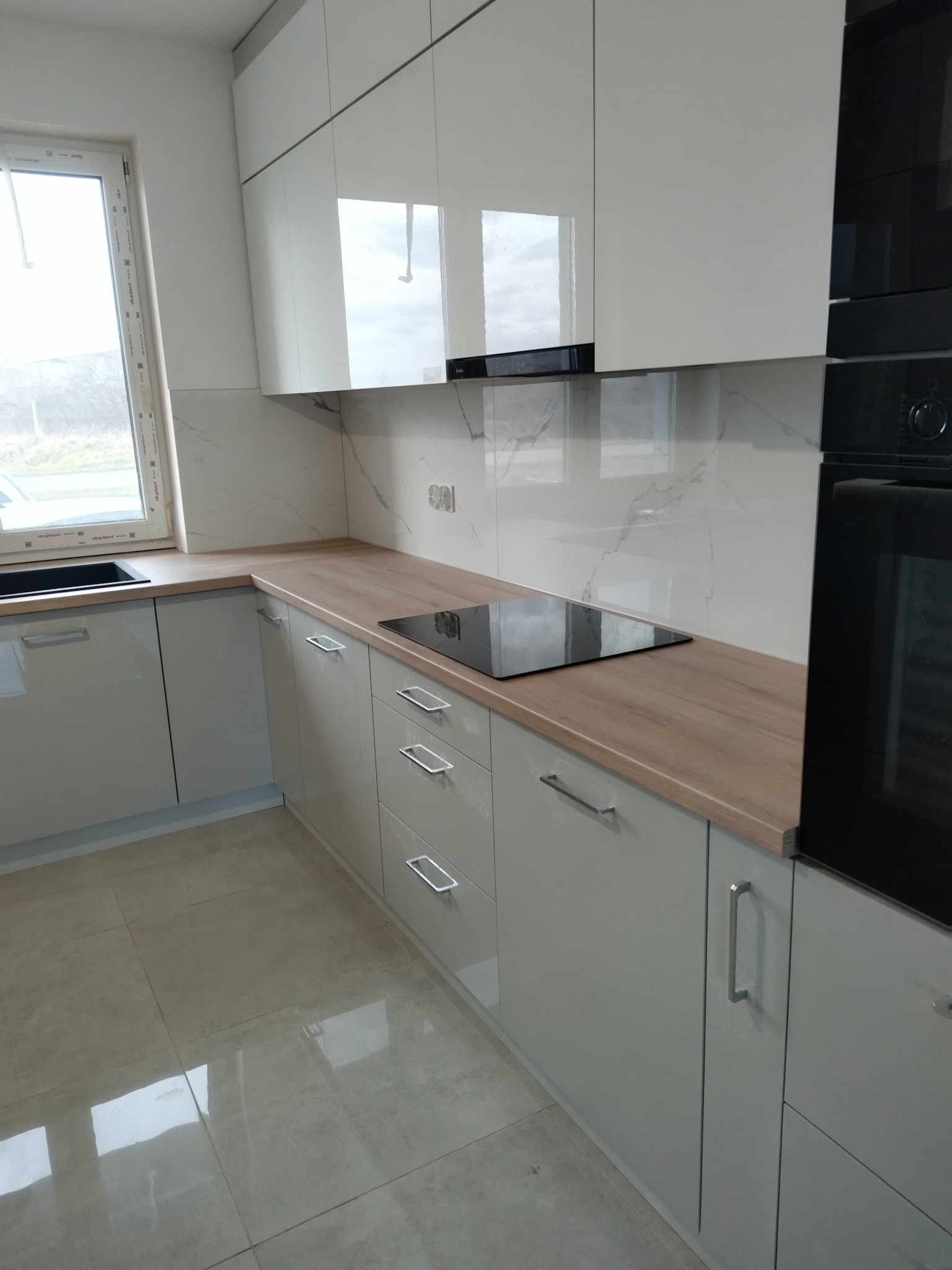 Nowoczesna kuchnia z błyszczącymi, szarymi szafkami, drewnianym blatem i płytą indukcyjną, wykończona marmurową mozaiką ścienną.