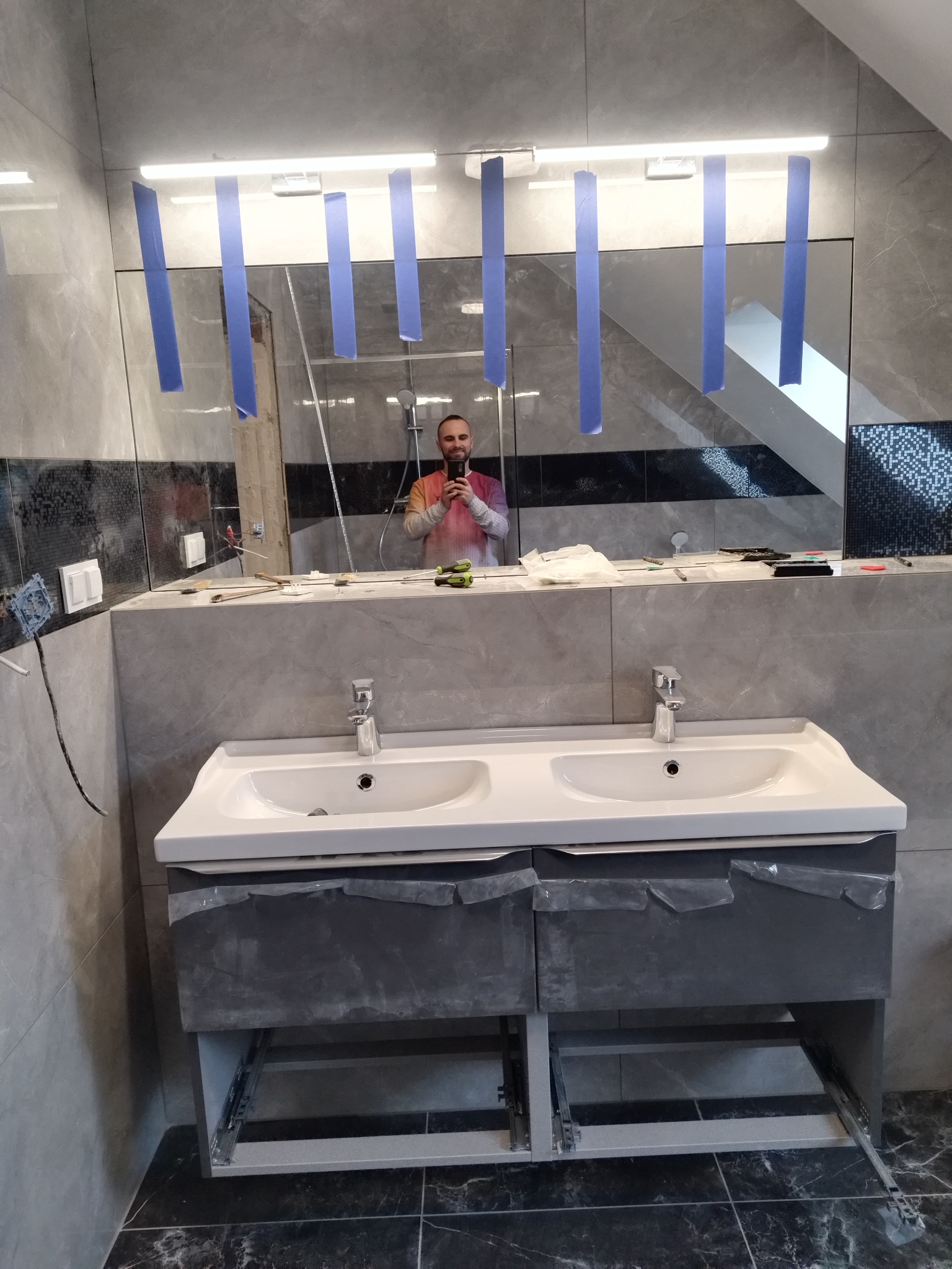 Podwójna umywalka z szarą szafką w trakcie montażu w łazience z szarymi płytkami, widoczne narzędzia i odbicie osoby robiącej zdjęcie w lustrze zabezpieczonym niebieską taśmą.