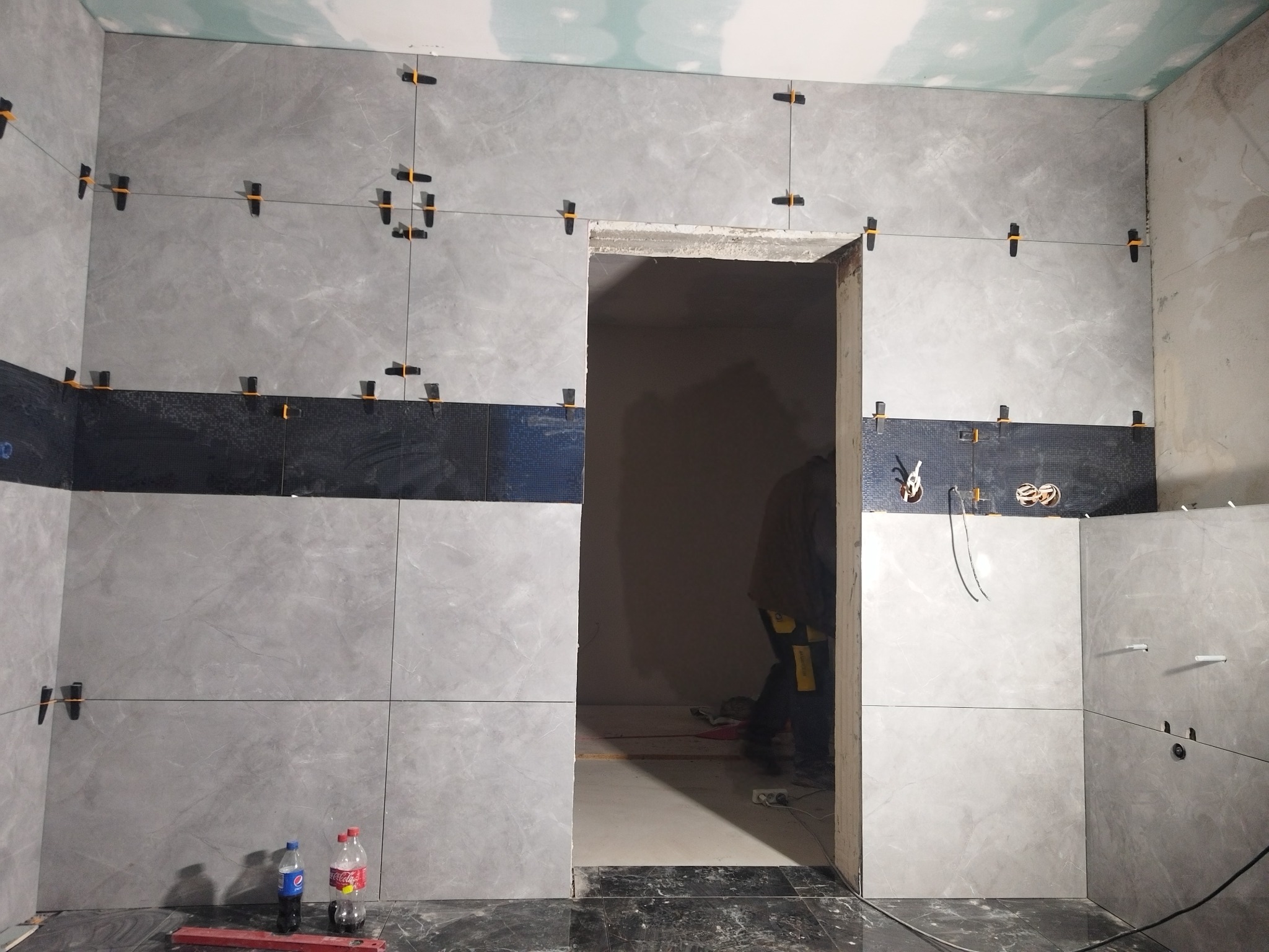 Szare płytki imitujące marmur, ułożone na ścianie z użyciem systemu poziomowania, z czarnym pasem dekoracyjnym na wysokości wzroku, widoczne butelki z napojami na podłodze i osoba w tle w przejściu.