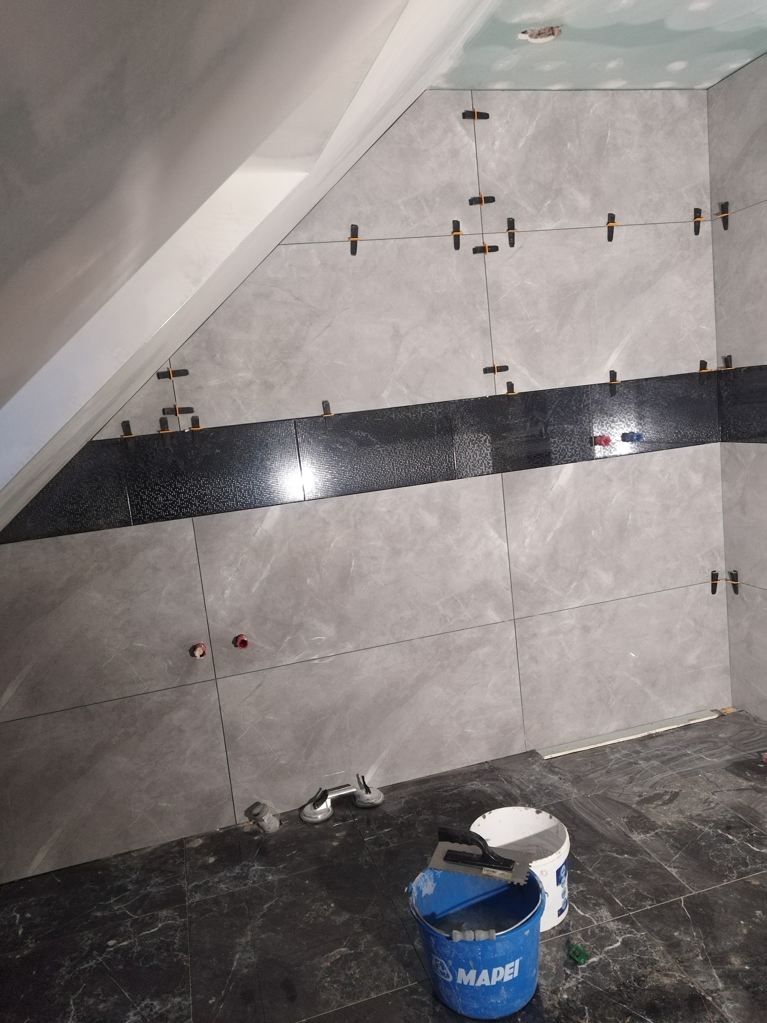 Ściana łazienki w trakcie układania płytek w szarym marmurkowym wzorze, z poziomym pasem czarnych płytek mozaikowych; widoczne krzyżyki dystansowe, przyssawki do płytek, wiadro z klejem Mapei...