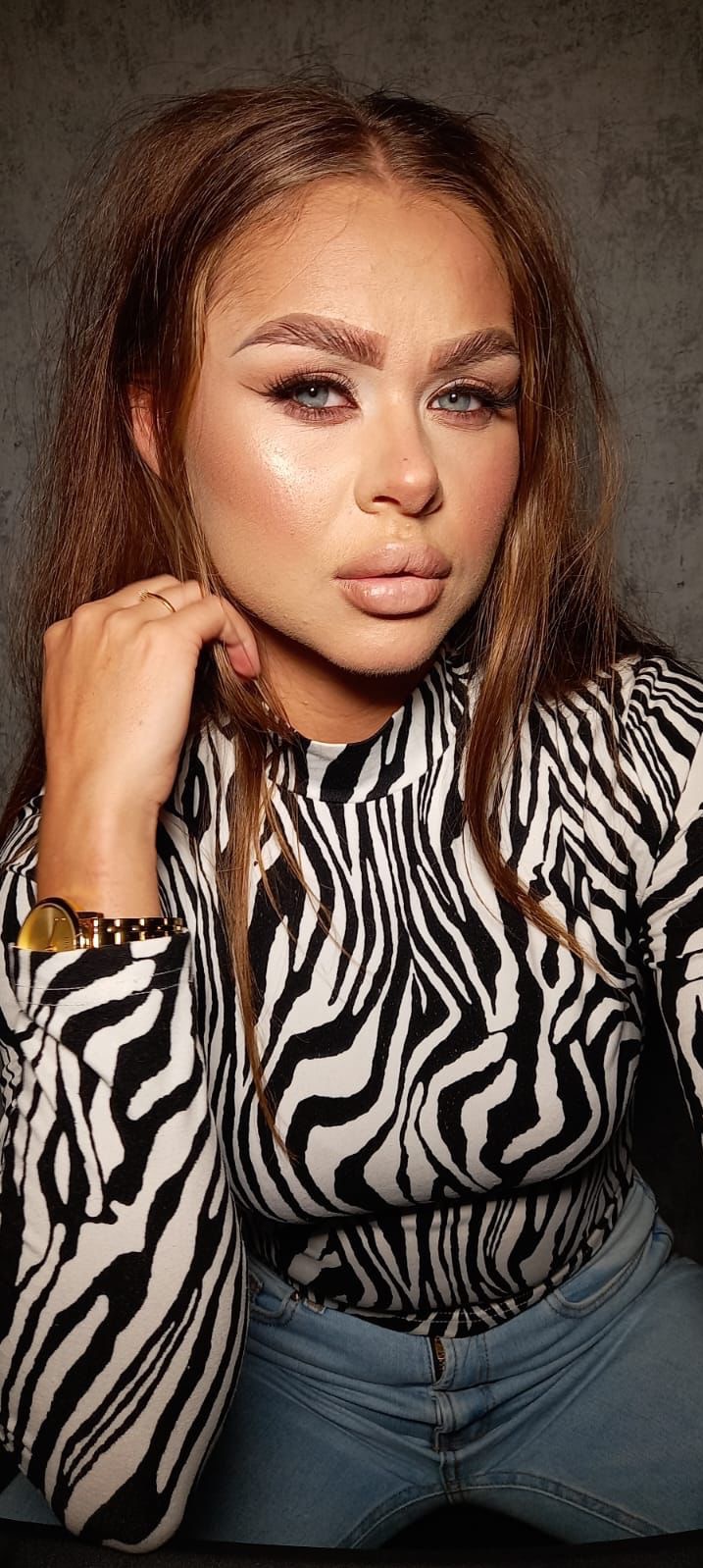 Portret kobiety z mocnym makijażem, w bluzce w zebrę i niebieskich jeansach, z złotym zegarkiem na ręce.