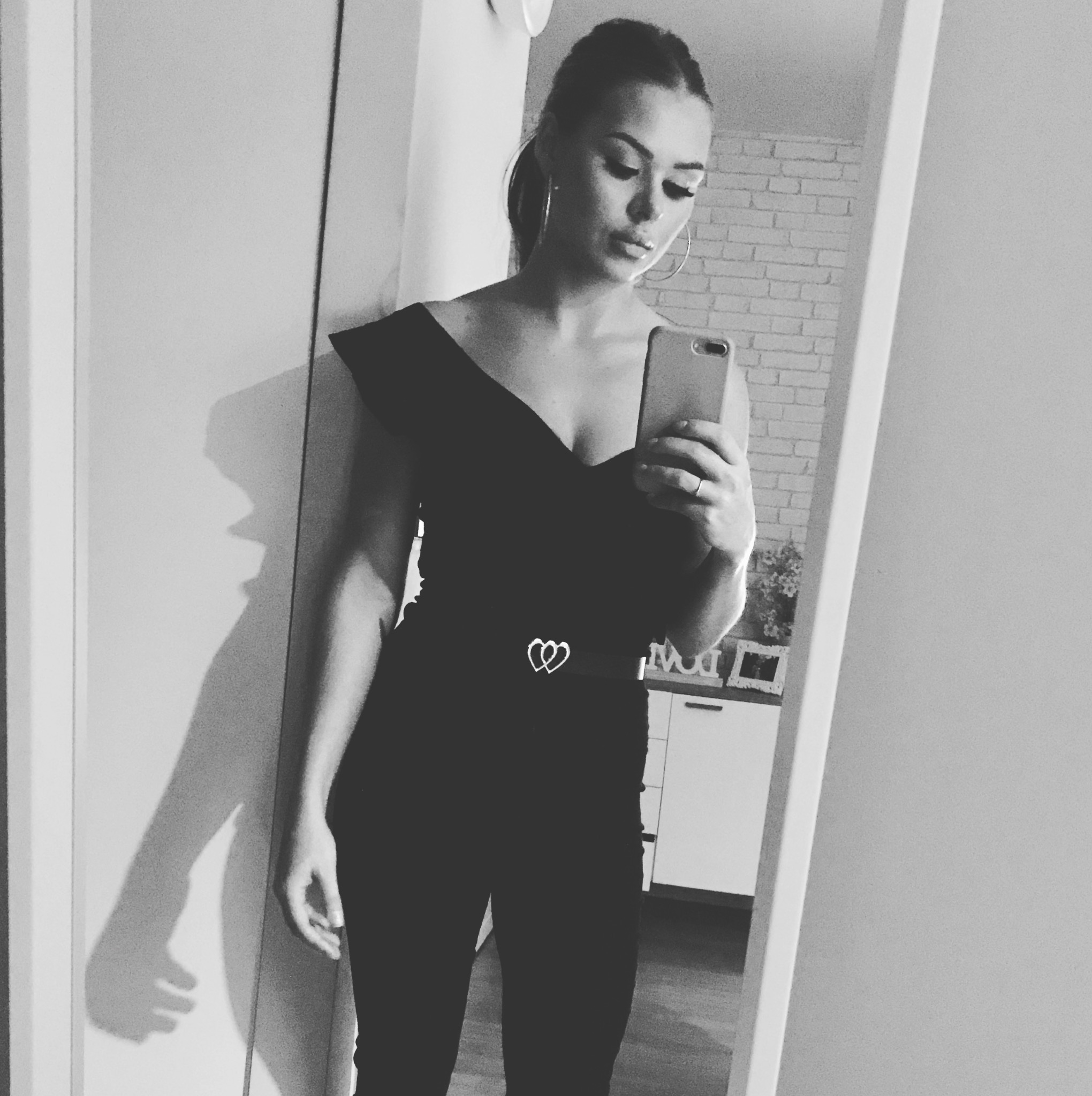 Kobieta w czarnym kombinezonie z odkrytymi ramionami pozuje do selfie w lustrze, widoczny pasek z klamrą w kształcie serc, jasny mur z cegły w tle.