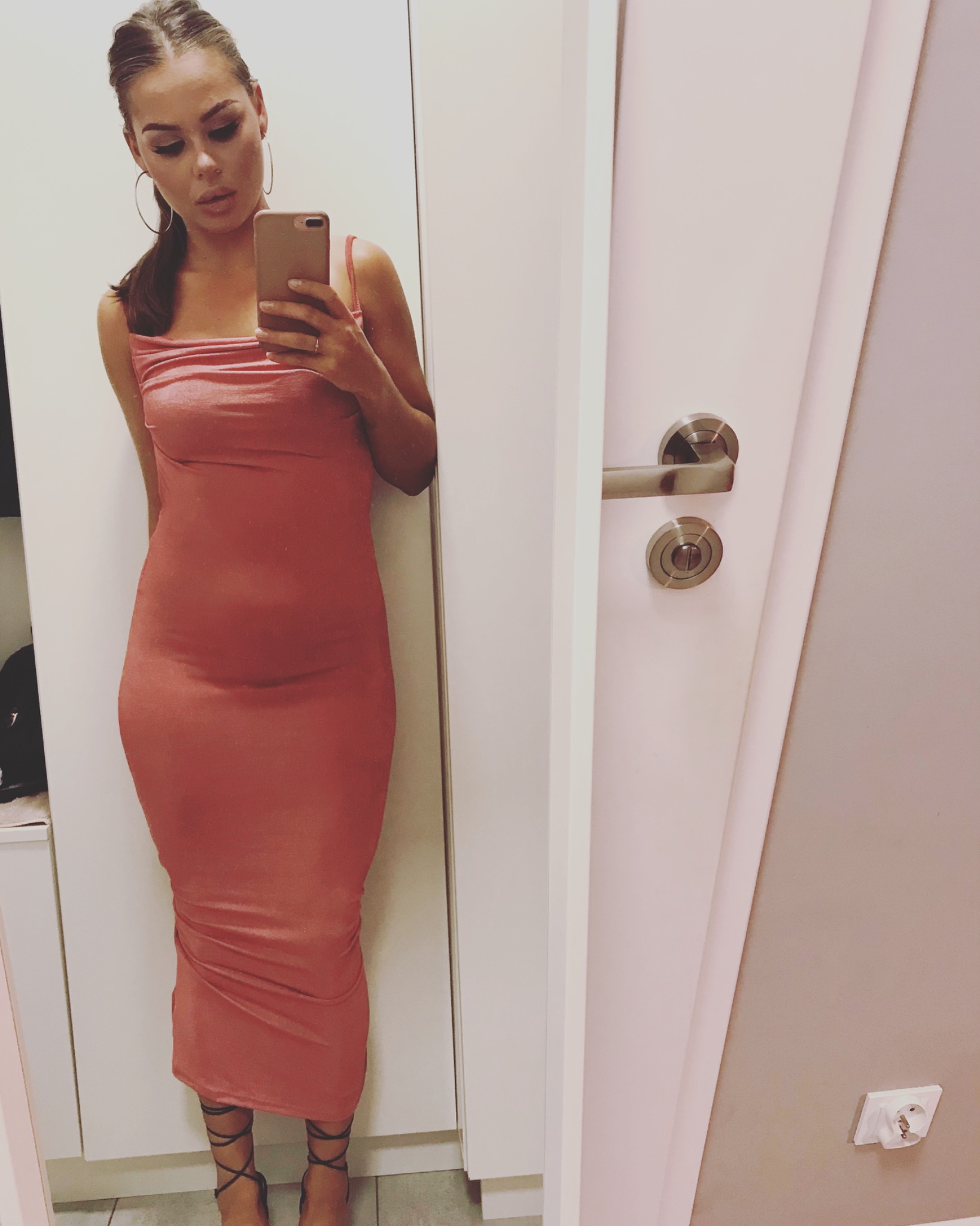 Kobieta w dopasowanej, koralowej sukience robi selfie w lustrze, widoczne czarne sandały na sznurowanie i srebrne kolczyki koła.