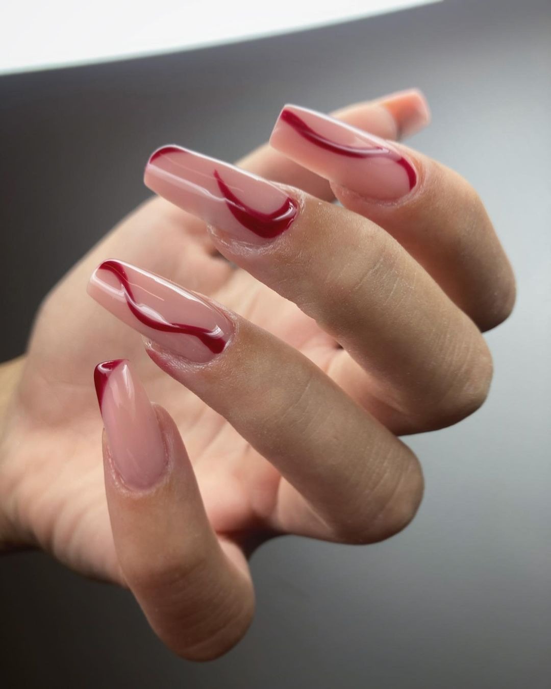 Dłoń z kwadratowymi paznokciami w kolorze nude z abstrakcyjnym wzorem w kolorze ciemnej czerwieni, wykonanym lakierem hybrydowym.