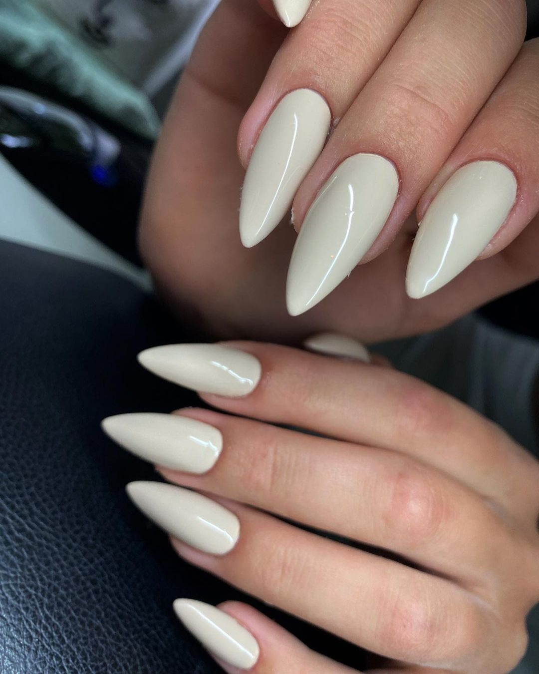 Dłonie z beżowymi, błyszczącymi paznokciami w kształcie szpiców na tle czarnej skórzanej powierzchni.