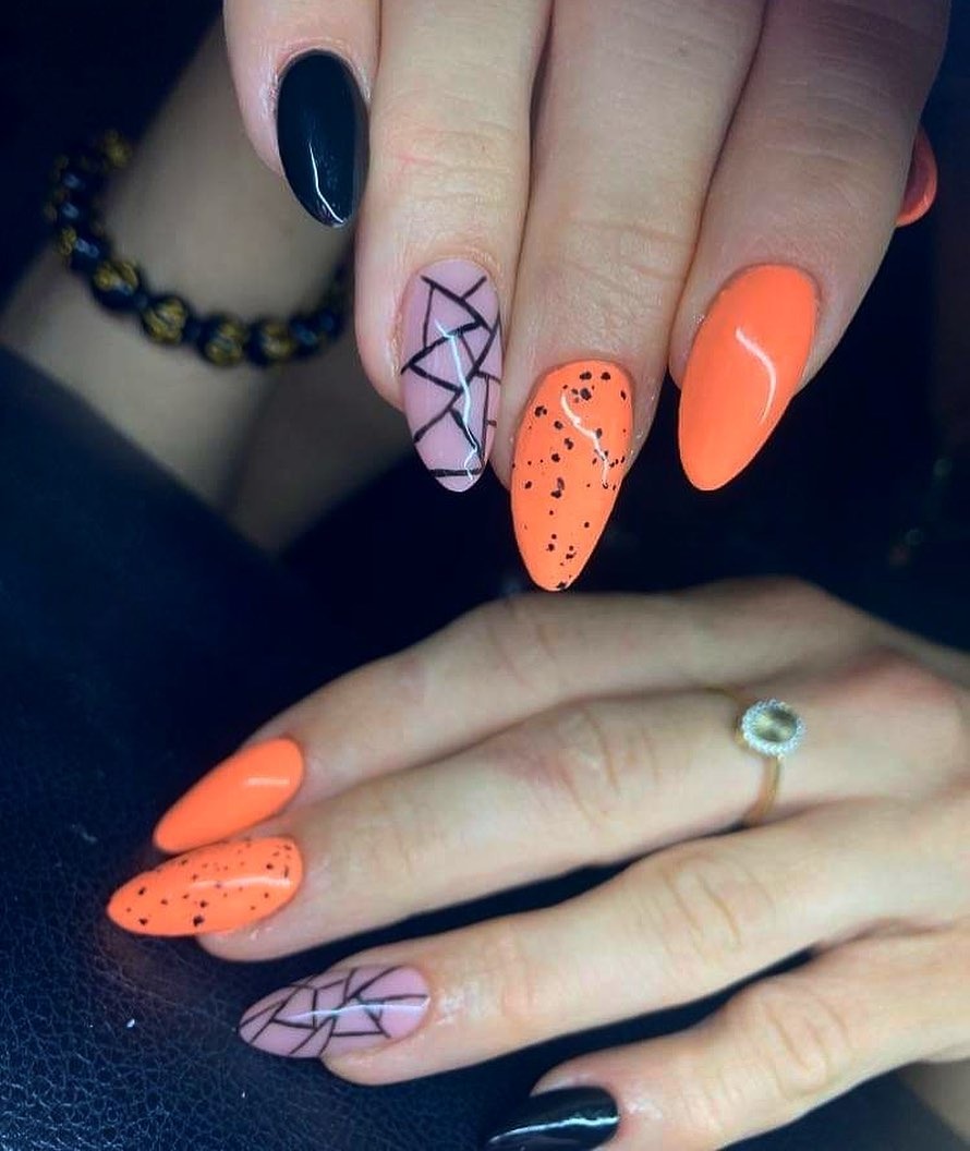 Dłonie z pomalowanymi paznokciami: jeden czarny, jeden różowy z czarnymi liniami, jeden pomarańczowy w czarne kropki i jeden gładki pomarańczowy, widoczny pierścionek na palcu