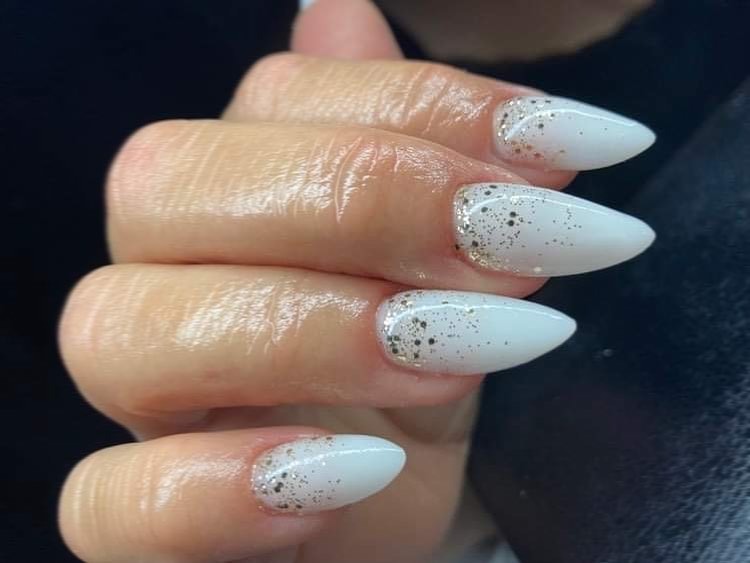 Dłoń z białymi, szpiczastymi paznokciami zdobionymi złotym brokatem u nasady, widoczne delikatne zmarszczki na skórze.