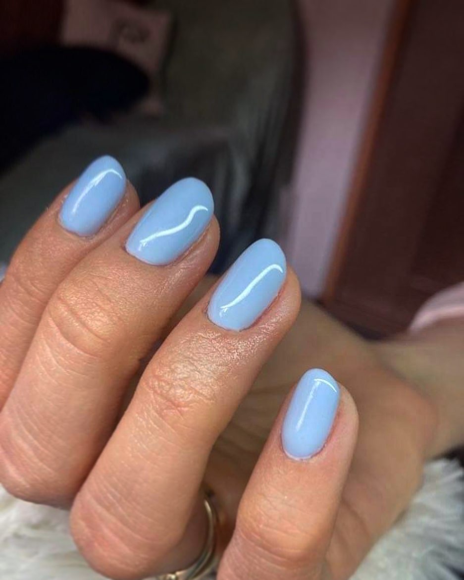 Dłoń z owalnymi paznokciami pomalowanymi na jasny, pastelowy błękit z delikatnym białym zdobieniem w kształcie fali na każdym paznokciu.