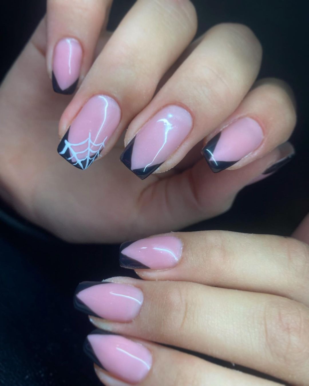 Dłonie z delikatnym, różowym manicure hybrydowym, czarne końcówki w kształcie litery V, na jednym paznokciu biała pajęczyna, błyszcząca powierzchnia.