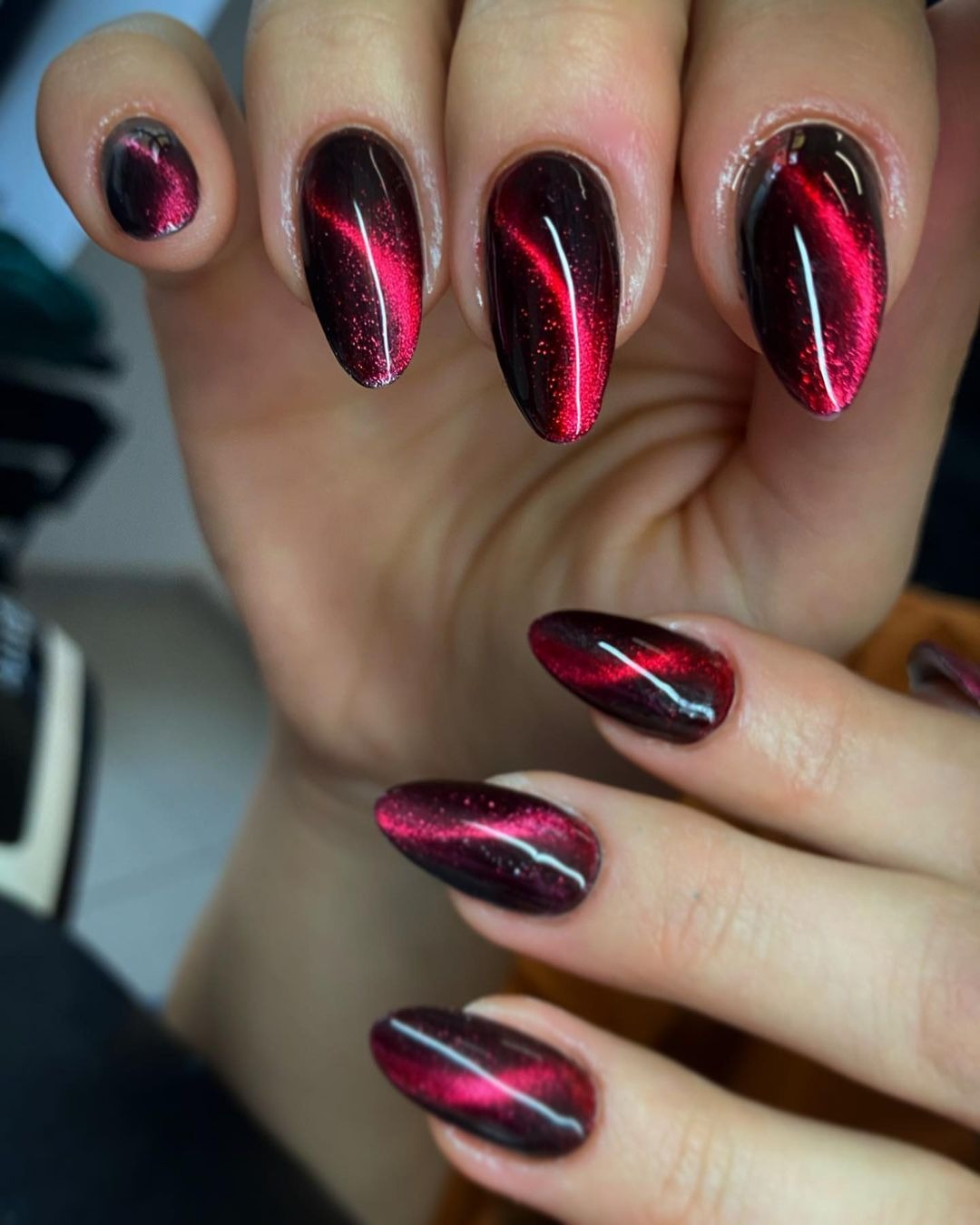 Dłoń z pomalowanymi na ciemny, bordowy kolor paznokciami w kształcie migdała z efektem kociego oka, gdzie czerwona linia światła biegnie przez środek każdego paznokcia.