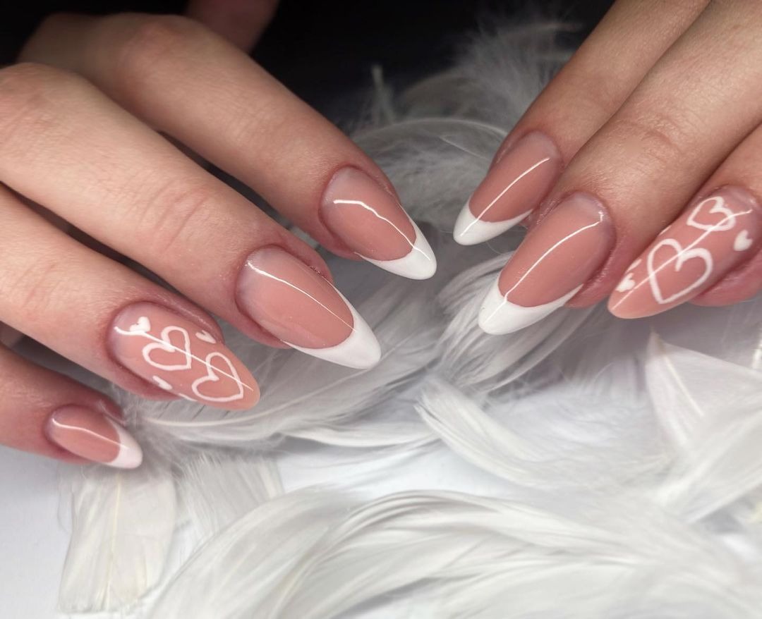 Dłonie z owalnymi paznokciami w stylu french z białymi końcówkami i wzorem białych serc na beżowym tle, ułożone na białych piórach.