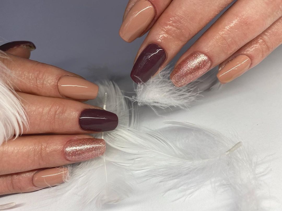 Dłonie z manicure hybrydowym: beżowy, bordowy i brokatowy lakier na tle białego piórka.