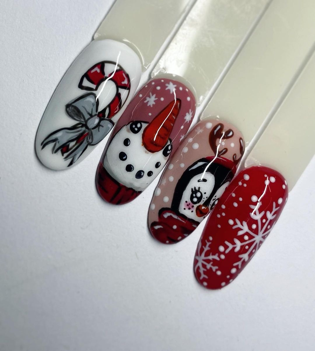 Zbliżenie na cztery tipsy z zimowymi motywami: lizak z kokardą, bałwan, pingwin w czapce i czerwony paznokieć ze śnieżynką, na białym tle.