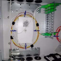 Wnętrze szafki telekomunikacyjnej z uporządkowanymi kablami światłowodowymi, zakończonymi zielonymi złączami i przymocowanymi niebieskimi uchwytami, widoczna etykieta ostrzegawcza o kablu...