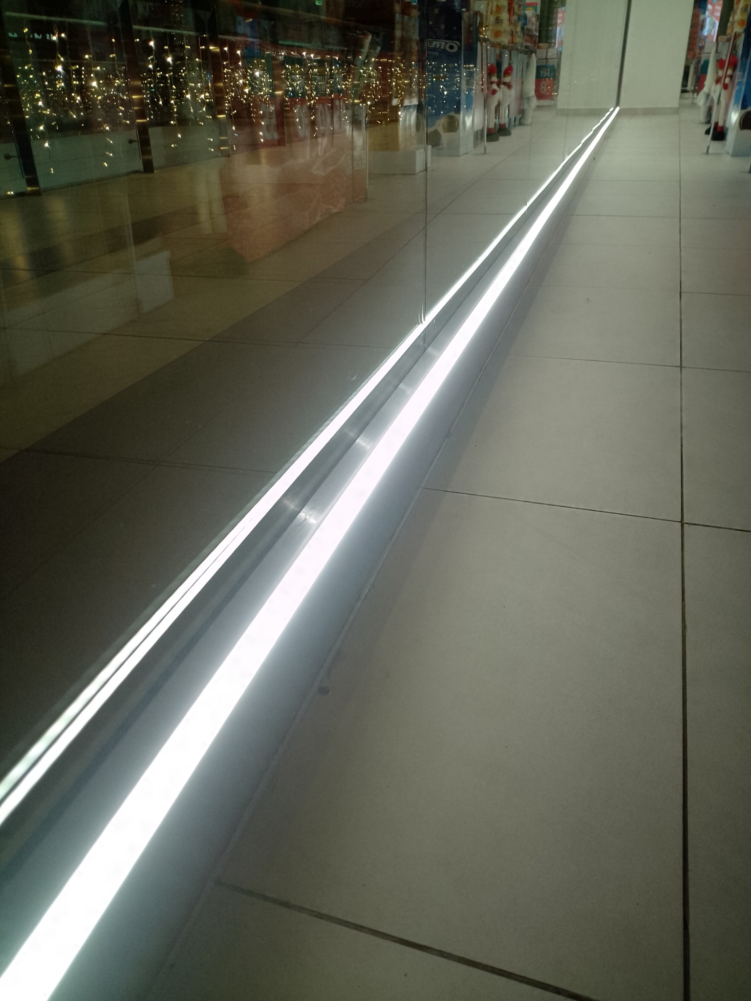 Wpuszczone w podłogę liniowe oświetlenie LED wzdłuż witryny sklepowej, odbijające światła i towary wewnątrz.