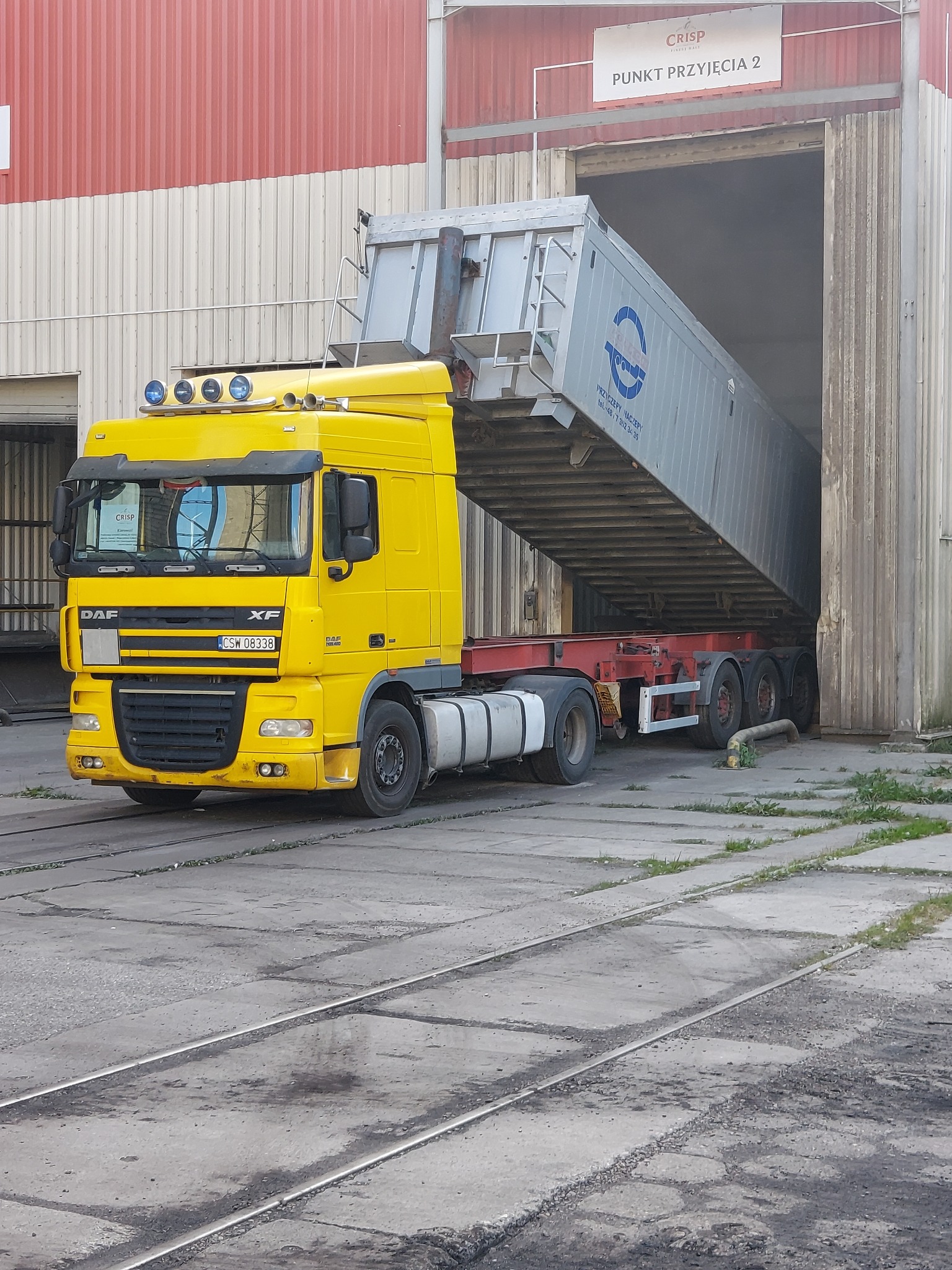 Żółta ciężarówka DAF XF z naczepą wyładowuje materiał do magazynu 'Punkt Przyjęcia 2', widoczne tory kolejowe w posadzce, logo na naczepie.