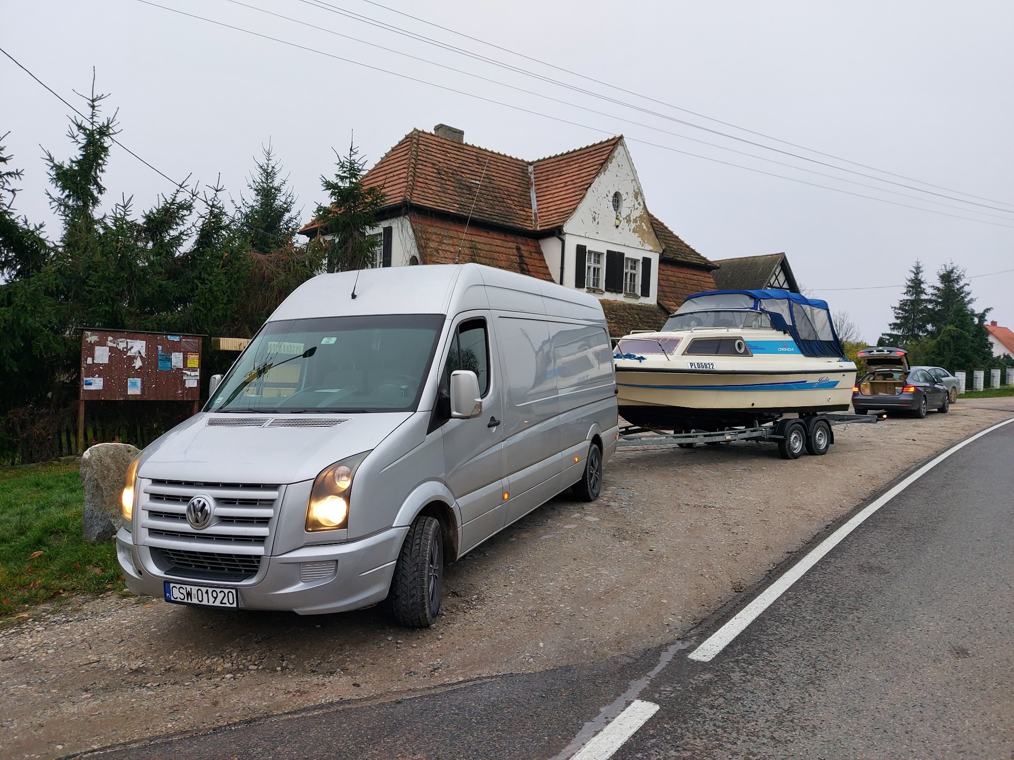 Srebrny van Volkswagen ciągnie przyczepę z łodzią na tle wiejskiego krajobrazu z domem o czerwonym dachu i tablicą ogłoszeń.