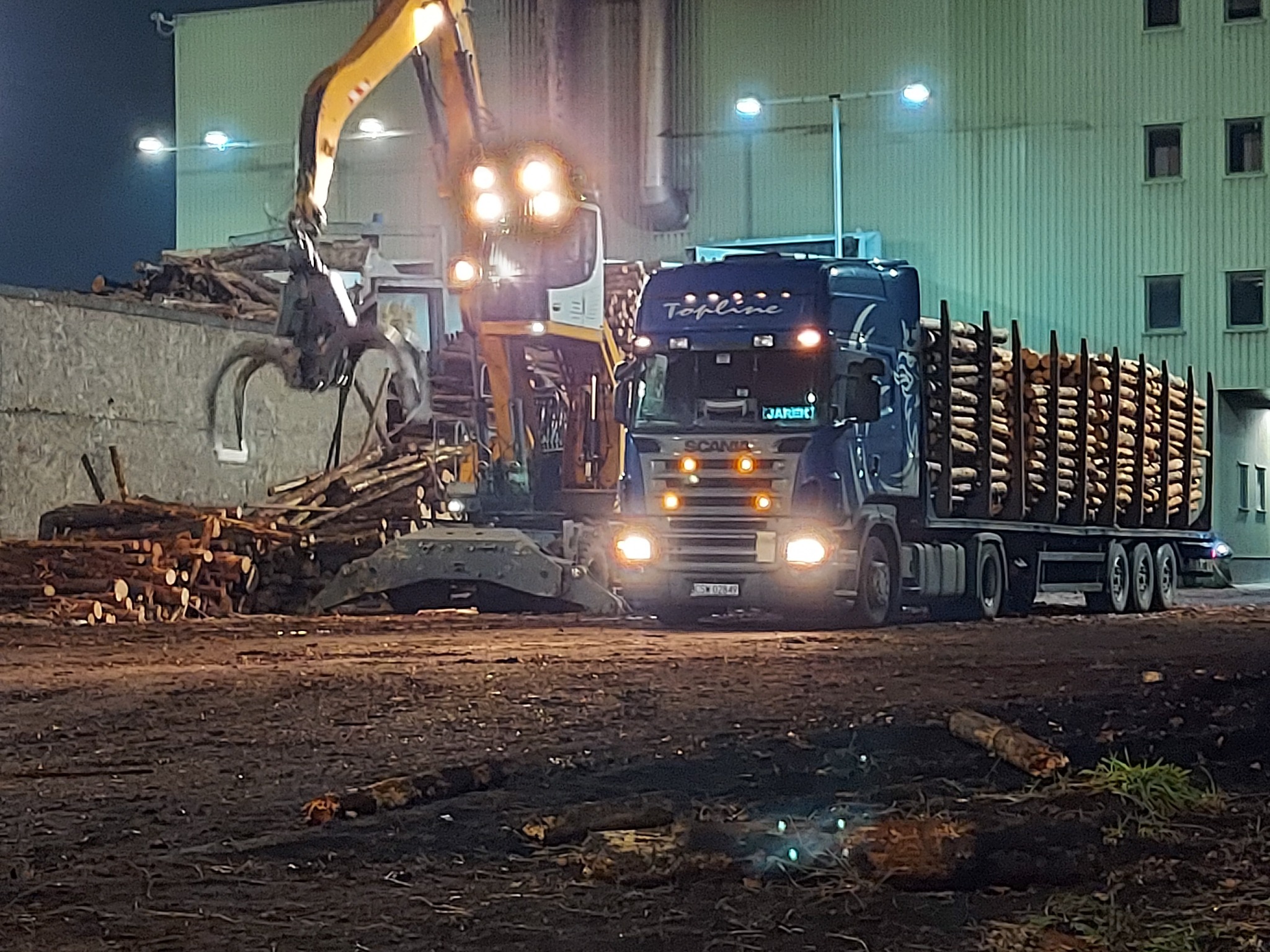 Ciężarówka Scania z niebieską kabiną Topline i załadowanymi kłodami drewna stoi obok koparki z chwytakiem, która układa kłody na stos, w tle budynek oświetlony lampami.