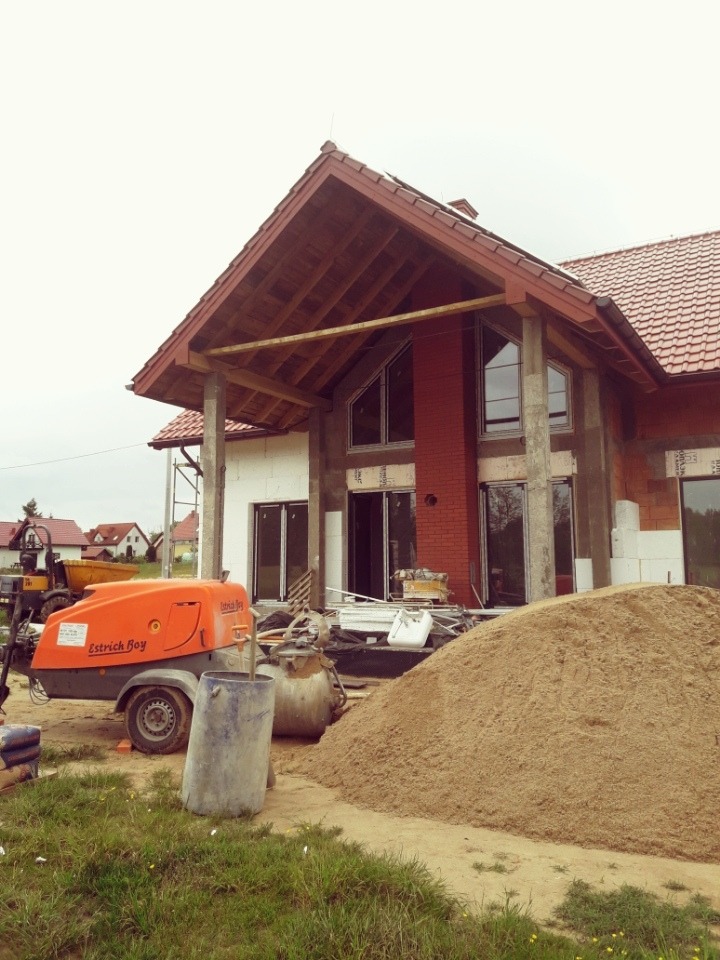 Budowa domu jednorodzinnego: widoczna maszyna do wylewania posadzek, sterta piasku i surowy stan wykończenia elewacji.