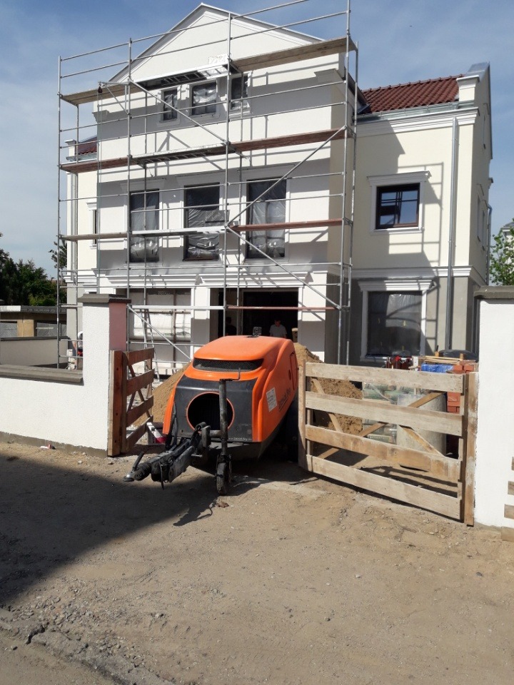 Pomarańczowa maszyna do wylewania posadzek stoi przed budynkiem w trakcie budowy, widoczne rusztowania i materiały budowlane.