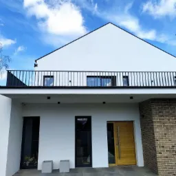 Nowoczesny dom z białą elewacją, czarną balustradą balkonową i drewnianymi drzwiami wejściowymi, sfotografowany w słoneczny dzień z błękitnym niebem i widocznymi chmurami.