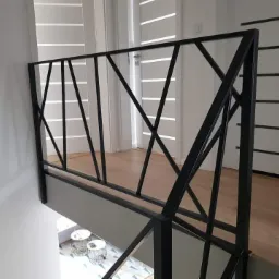 Nowoczesna czarna balustrada z geometrycznym wzorem na tle jasnego wnętrza z drewnianą podłogą i białymi drzwiami z poziomymi paskami.