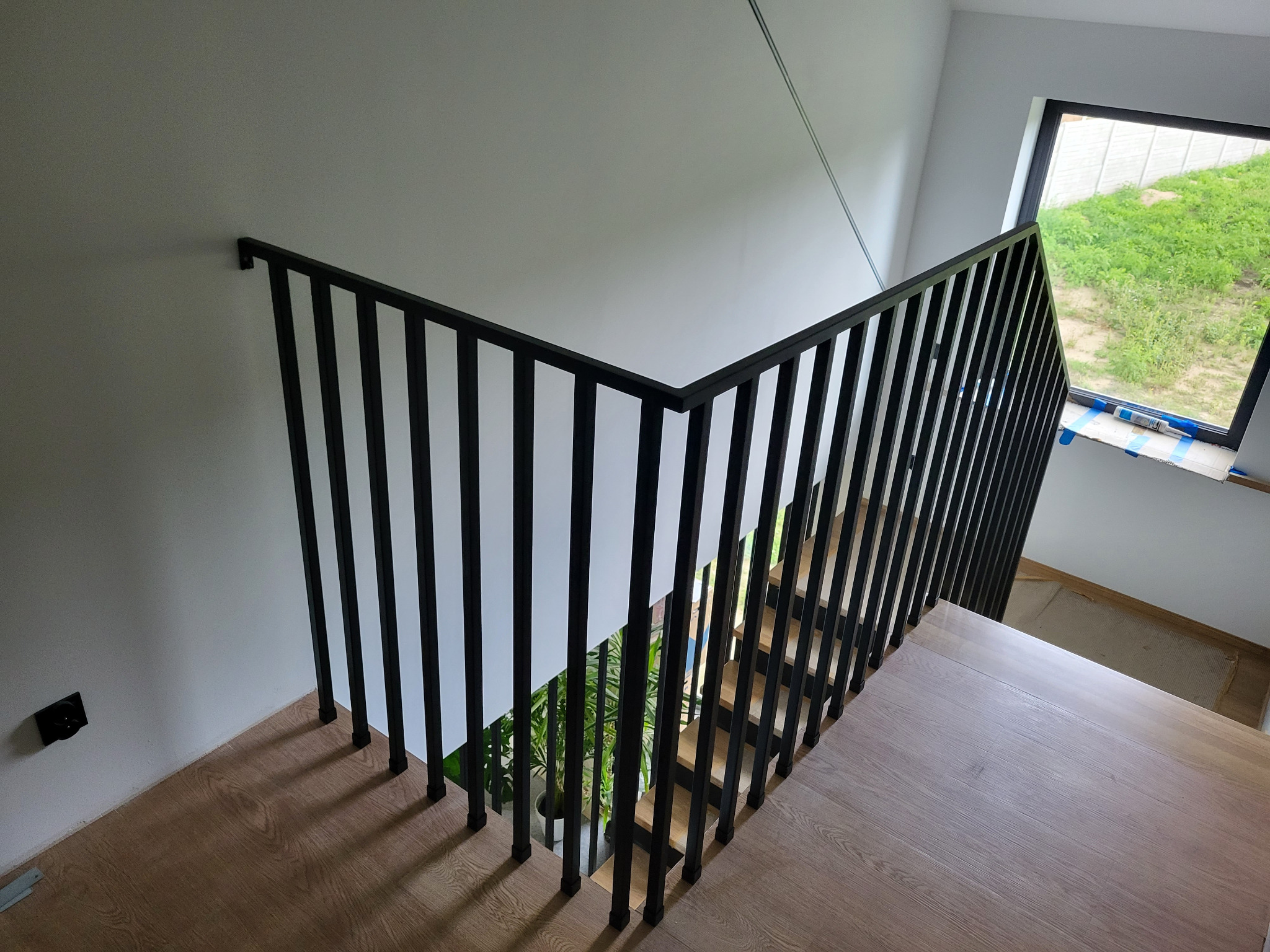 Nowoczesne schody z drewnianymi stopniami i czarną, pionową balustradą. Widok z góry na schody prowadzące w dół, z rośliną doniczkową widoczną przez szczeliny balustrady.