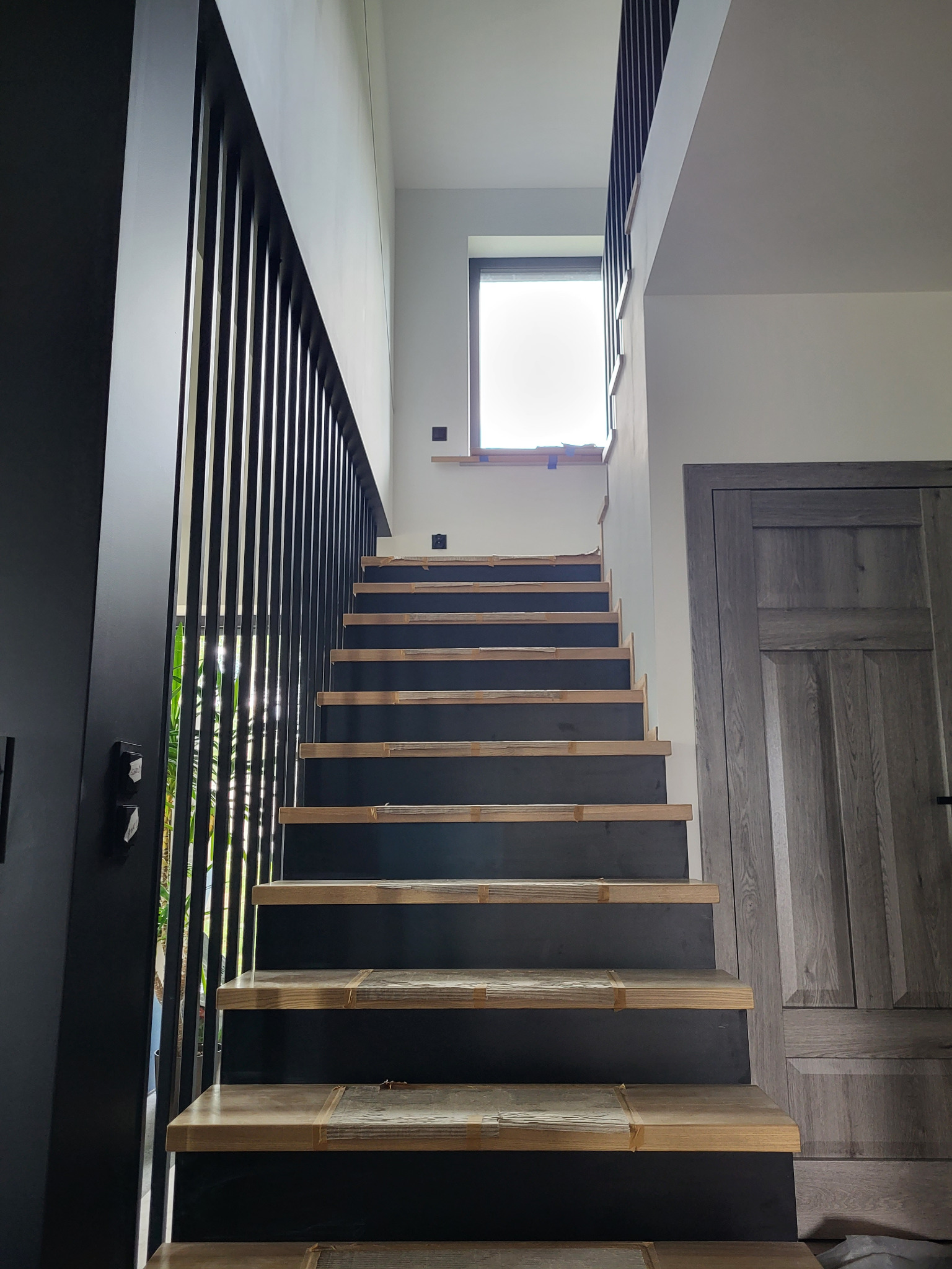 Wnętrze domu w trakcie wykańczania, widok na schody z drewnianymi stopniami zabezpieczonymi taśmą, czarne pionowe elementy balustrady, szare drzwi w tle i okno na poddaszu.