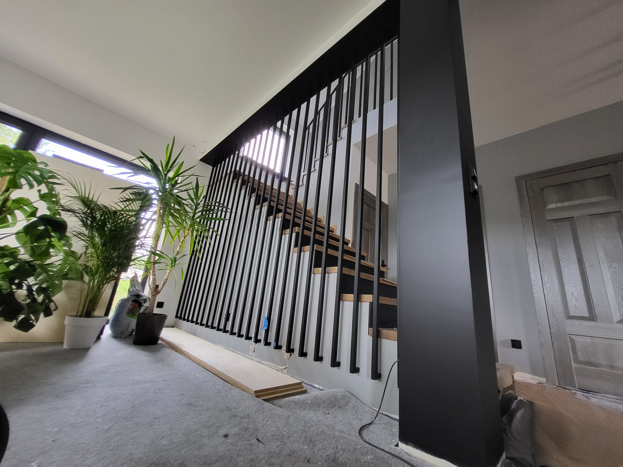 Nowoczesne schody z czarną, pionową konstrukcją zabezpieczającą, widoczne w surowym wnętrzu z roślinami doniczkowymi i przygotowanymi elementami podłogi.