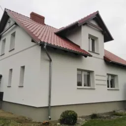 Nowoczesny dom jednorodzinny z białą elewacją, czerwoną dachówką i szarymi akcentami dekoracyjnymi na ścianach, widok z boku w pochmurny dzień.