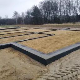 Przygotowany fundament pod budowę domu jednorodzinnego, widoczne szalunki wypełnione piaskiem oraz wystające rury kanalizacyjne.