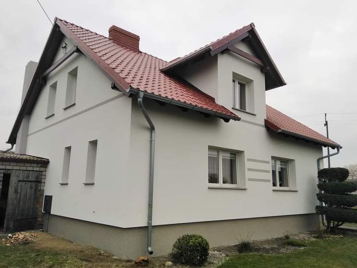 Nowoczesny dom jednorodzinny z białą elewacją, czerwoną dachówką i szarymi akcentami dekoracyjnymi na ścianach, widok z boku w pochmurny dzień.