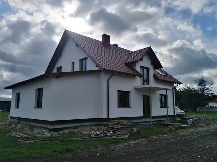 Świeżo wybudowany, dwukondygnacyjny dom z białymi ścianami i czerwoną dachówką, widoczny z boku, z paletami i materiałami budowlanymi na trawniku przed domem.