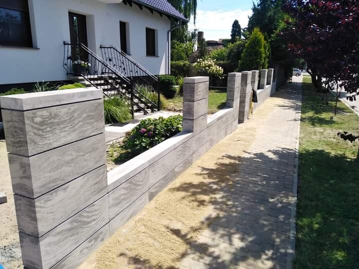 Nowoczesny ogrodzenie z betonowych bloczków o szarej, marmurkowej fakturze, oddzielające posesję od chodnika z kostki brukowej; widoczny dom z białymi ścianami i schodami z czarną balustradą.