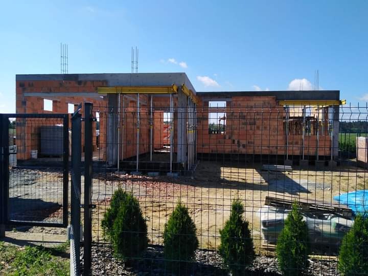 Budowa domu w stanie surowym otwartym, widok zza ogrodzenia panelowego i rzędu małych tuj, niebo błękitne z lekkimi chmurami.