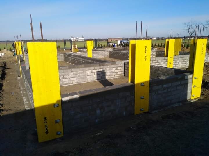 Wykop pod fundamenty domu w trakcie budowy, widoczne szalunki w kolorze żółtym oraz fragmenty muru z bloczków betonowych. W tle zielone pola i zabudowania.